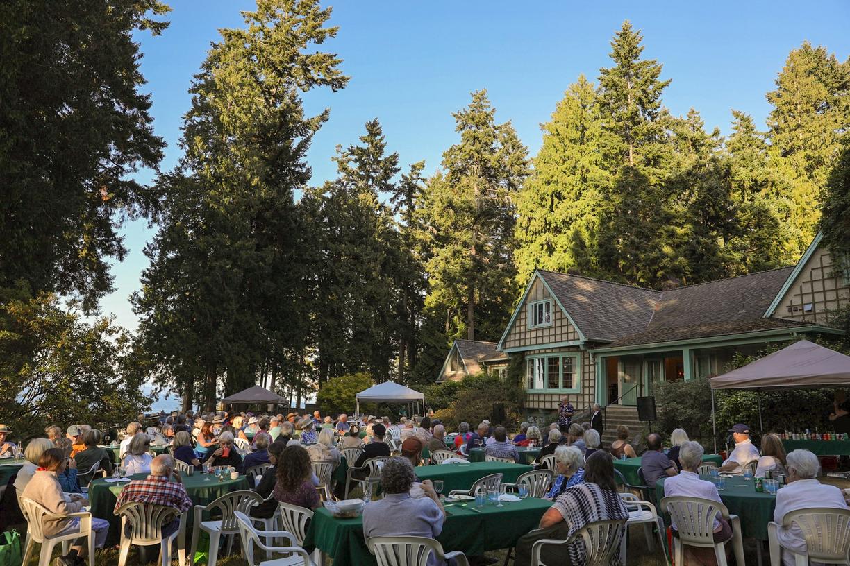 hundreds of people sitting around tables set out on a lawn with a cottage in the background