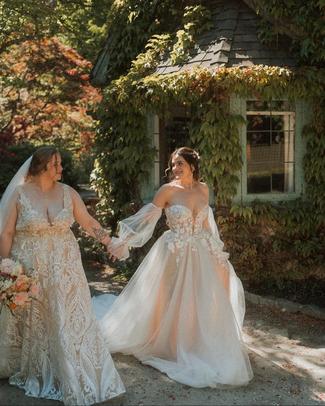 brides holding hands in front of vine covered garden cottage.